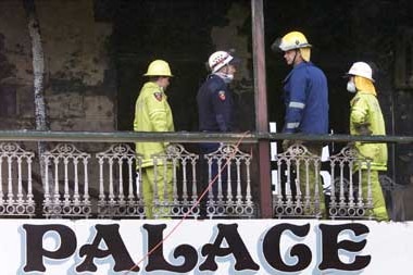 Four emergency service workers wear hard hats as they stand on the top verandah of burnt-out hotel