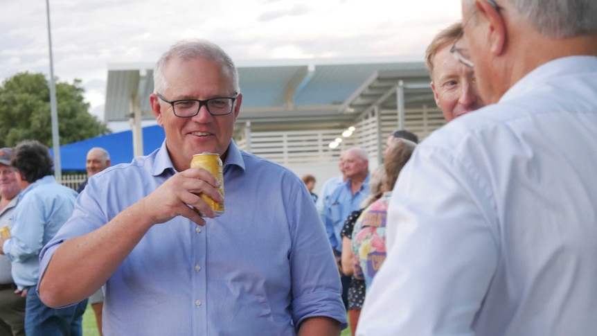 Prime Minister stands in a group, smiling and looking at camera while holding a can of drink.