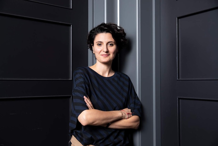 Tallish, lean woman with short dark wavy hair, wearing long-sleeved top with black and navy diagonal stripes, arms folded.