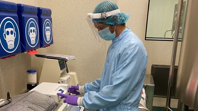 A man in medical scrubs, a mask and hair-net inspects medical vials.