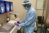 A man in medical scrubs, a mask and hair-net inspects medical vials.
