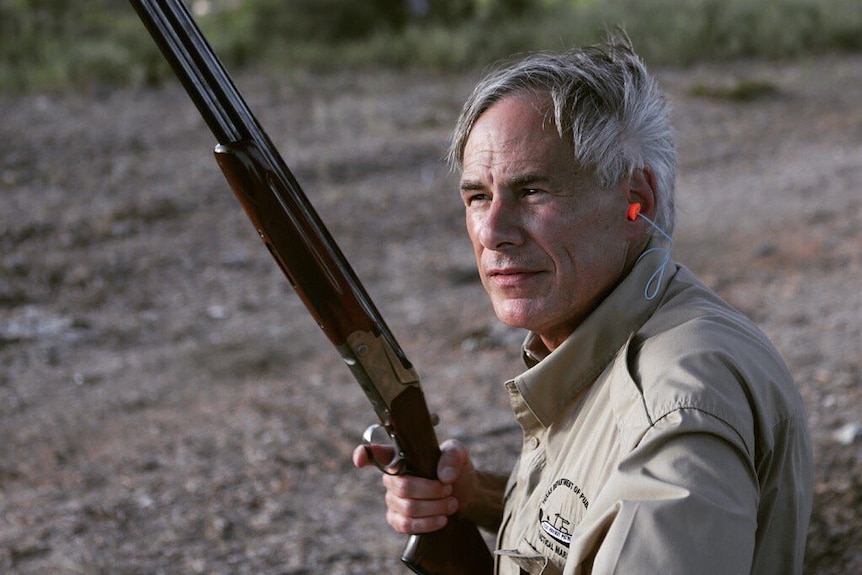 A man with silver hair, dressed in a khaki shirt, looks off in the distance while holding a rifle