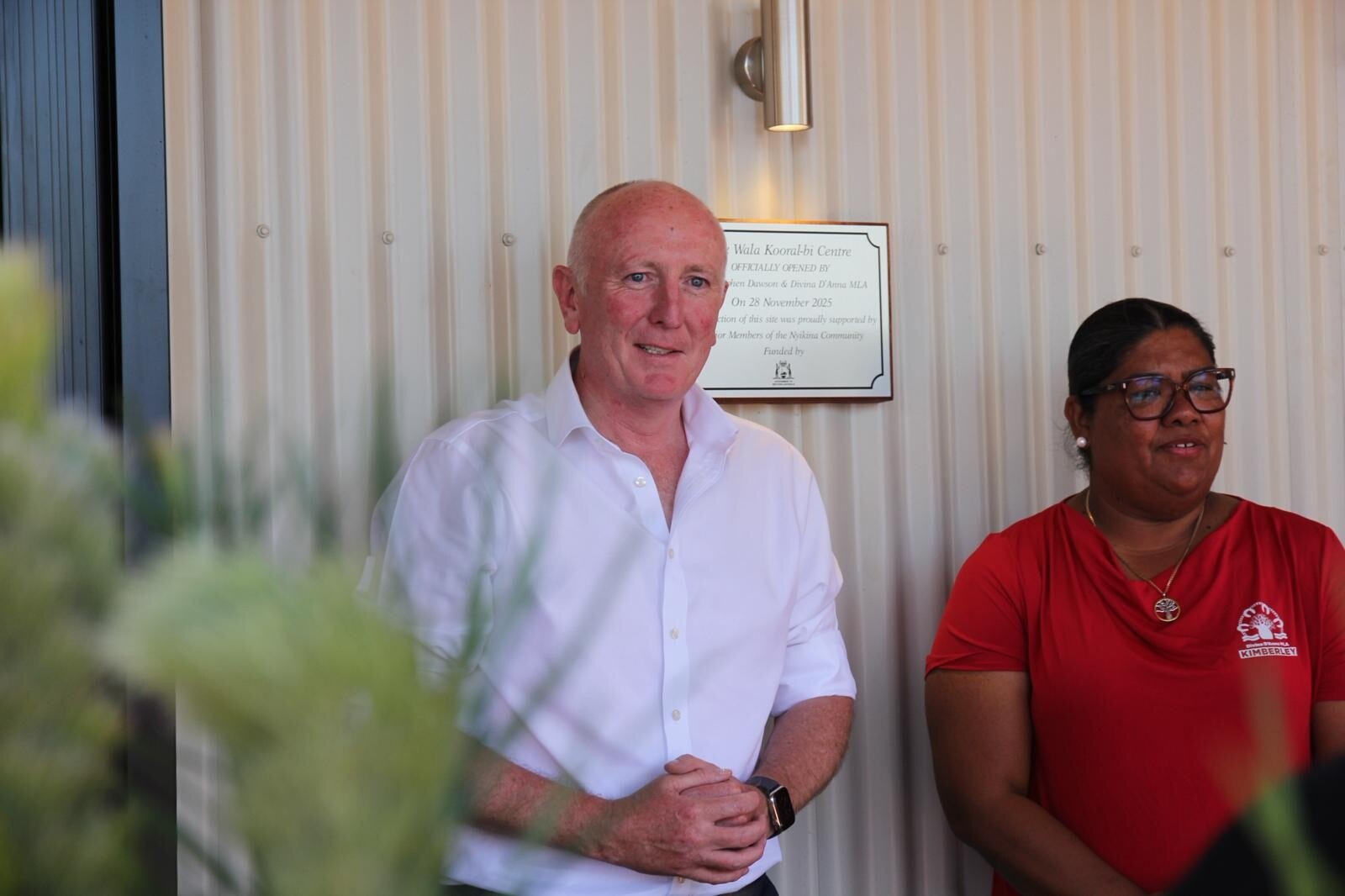 Un hombre de mediana edad con camisa de negocios está de pie con una mujer de cabello oscuro frente a un edificio de hojalata.