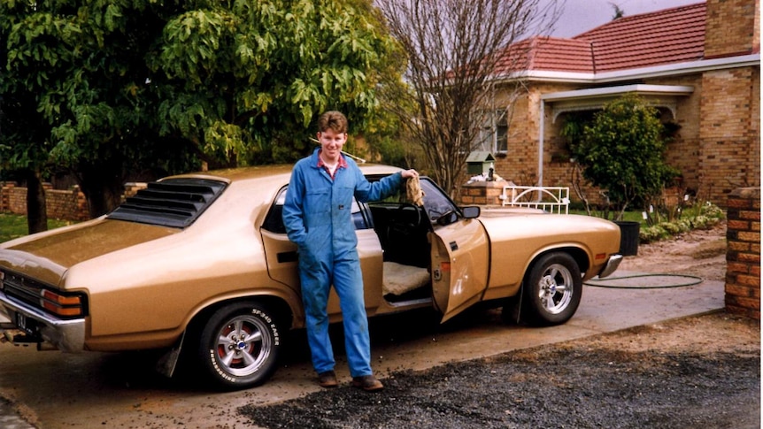 A young man in a boiler suit poses in a driveway leaning on the open door of a retro-style car.