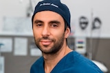 A man wearing a surgeon's scrubs and a hat with his name embroidered on it looks directly into the camera.