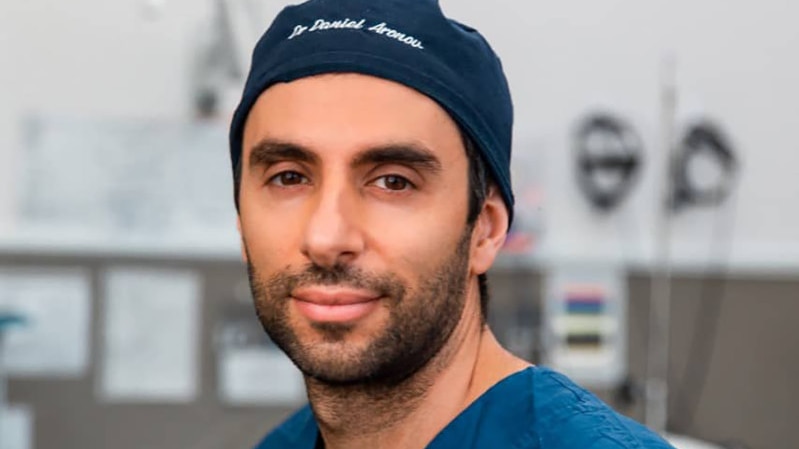 A man wearing a surgeon's scrubs and a hat with his name embroidered on it looks directly into the camera.