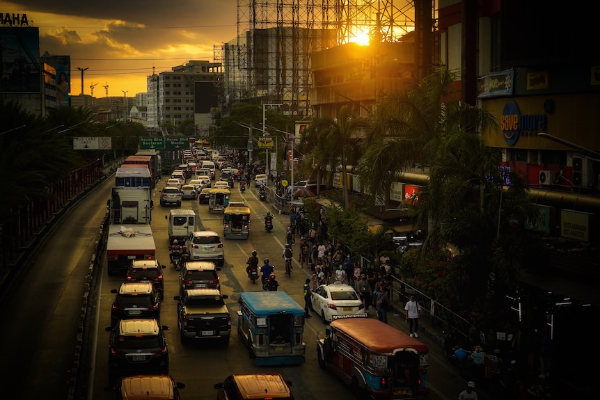 Roads, buses and trucks line a busy road seen from a bridge above.  The sun is setting in the background