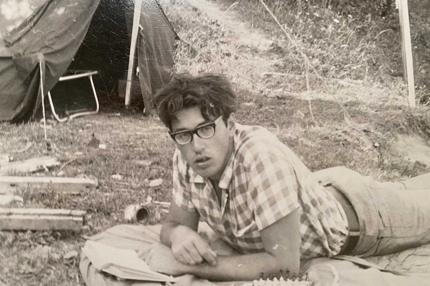 Mike lies on the ground reading a book in a black and white photo. He looks up at the camera, behind him is a tent.