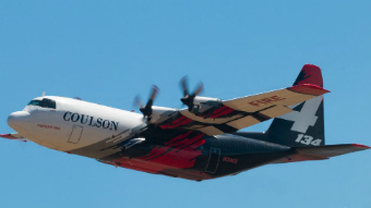 A water bomber is seen with blue skies overhead. Its propellers are active, half of the plane is white and the back half is navy