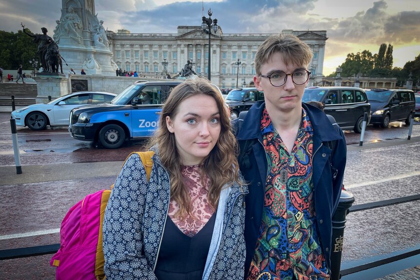 A young woman and a man in glasses stand outside Buckingham Palace 