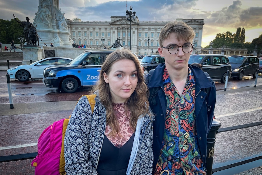 A young woman and a man in glasses stand outside Buckingham Palace 