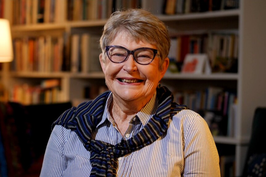 Smiling mature woman in front of a dimly lit home library, short grey hair and glasses with a scarf draped around her shoulders