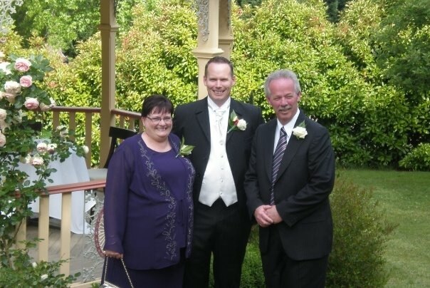 Ben Curtis, smiling and wearing a wedding suit, stands with his parents in a garden.