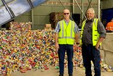 Two men stand in front of a pile of recyclable cans at the Cherbourg Material Recovery Centre