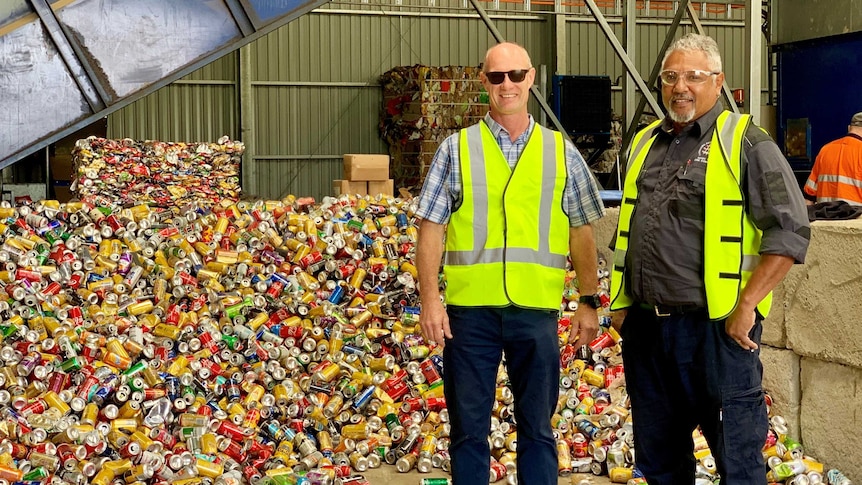 Two men stand in front of a pile of recyclable cans at the Cherbourg Material Recovery Centre.