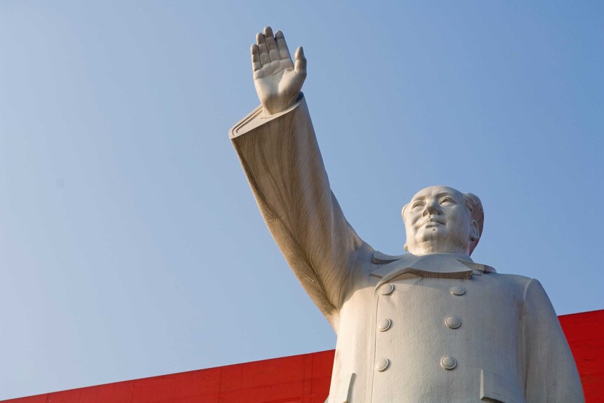 A statue of Mao Zedong with arm raised and blue sky behind.
