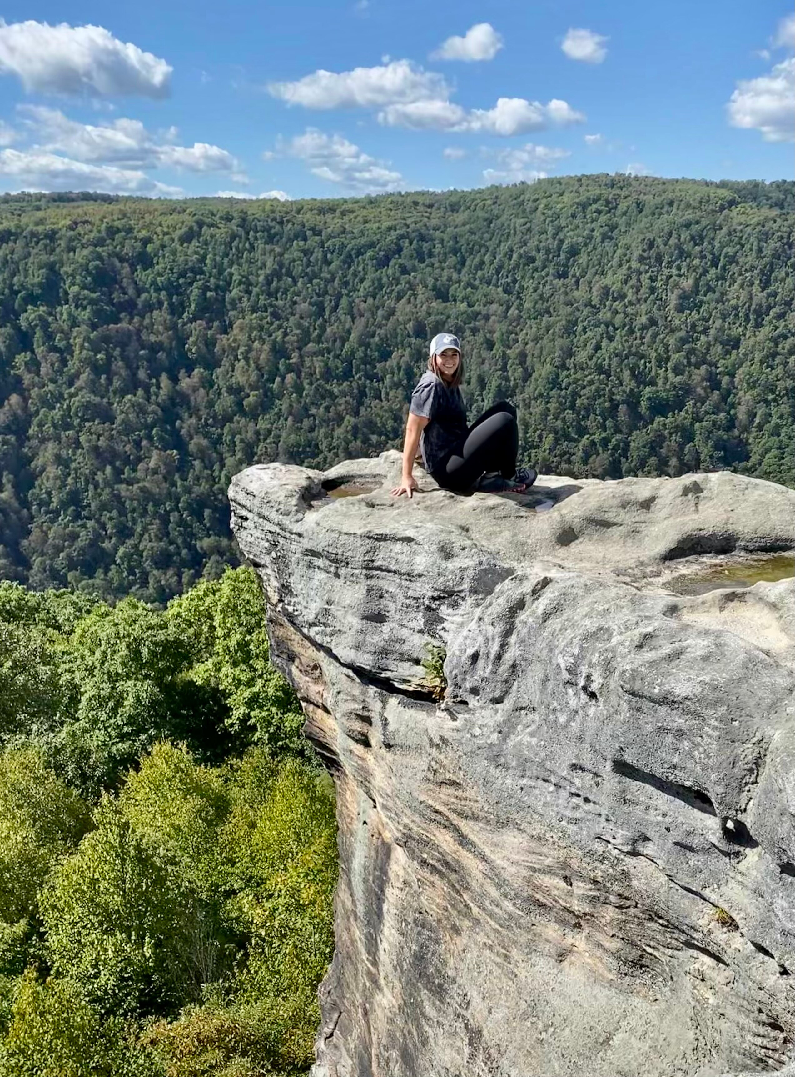 A young woman in a cap sits on a rock cliff in a forest 