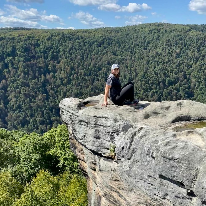 A young woman in a cap sits on a rock cliff in a forest 