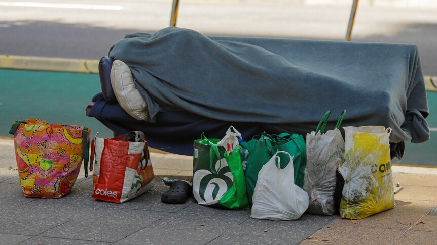 A homeless person is covered in a blanket while sleeping on a bench, their belongings in bags on the ground