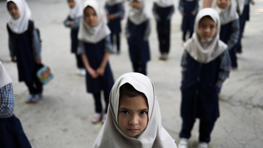 Afghan school girls stand outside wearing uniforms and headscarves.