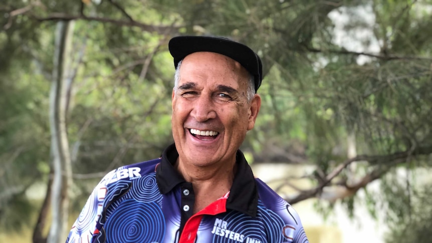 A smiling man in a blue top and a black cap laughs with trees behind him.