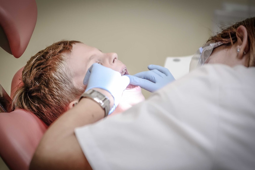 A young blonde boy is sitting in the dentists chair while the dentist examines his mouth