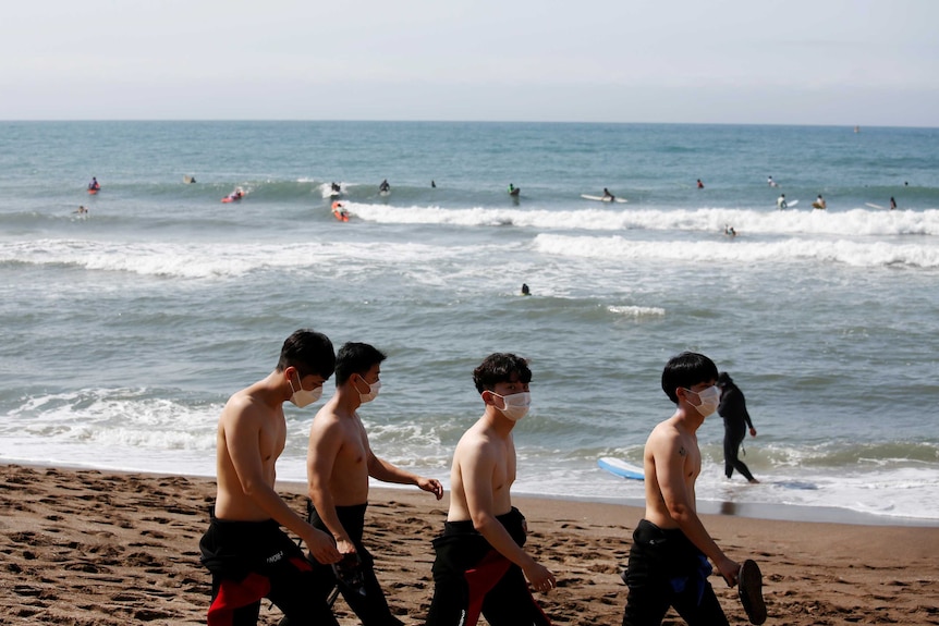 Four men wearing face masks walk on a beach as a number of people on surfboards swim behind them in the ocean