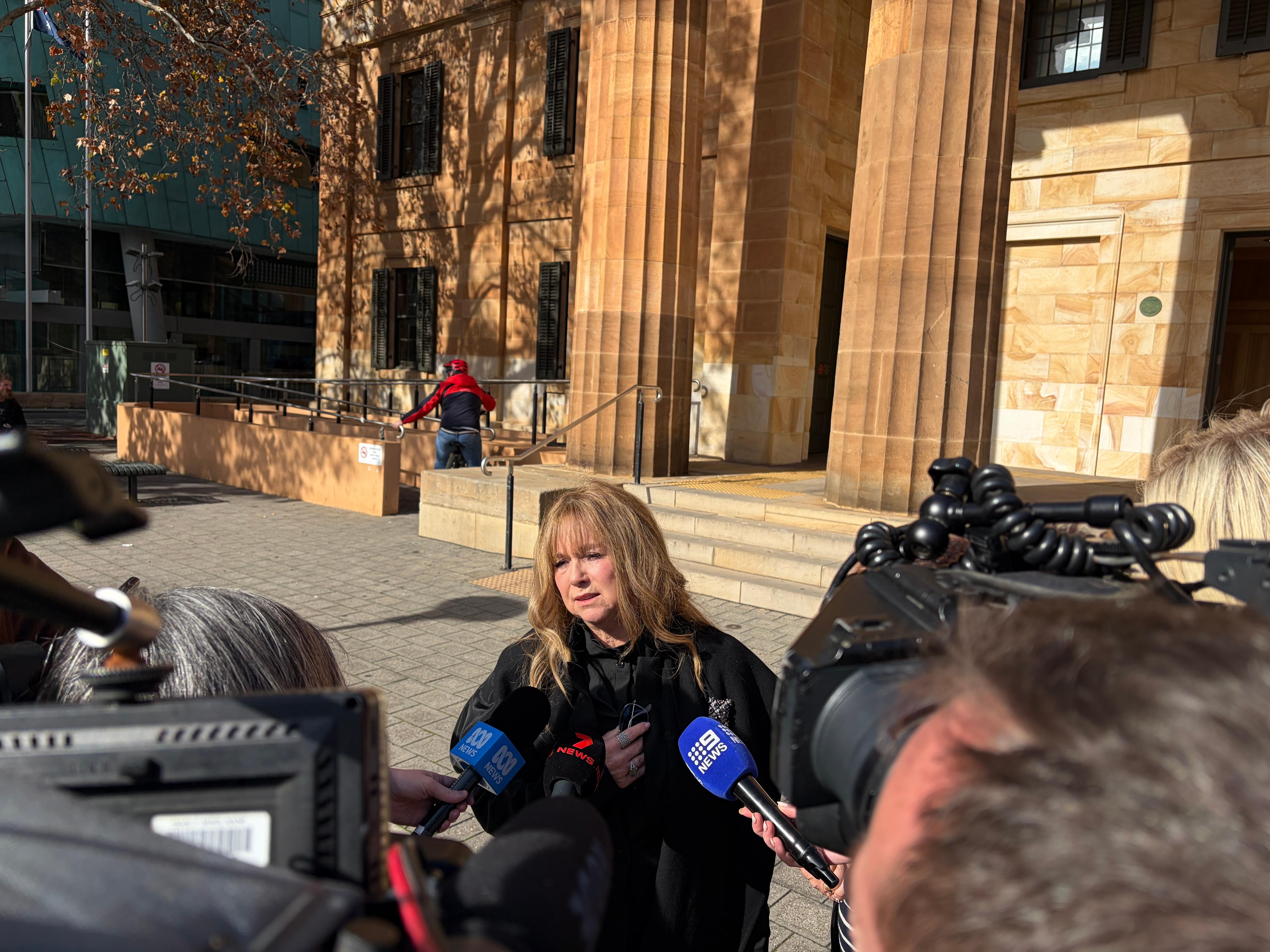 A blonde woman speaks to reporters holding microphones outside a stone building