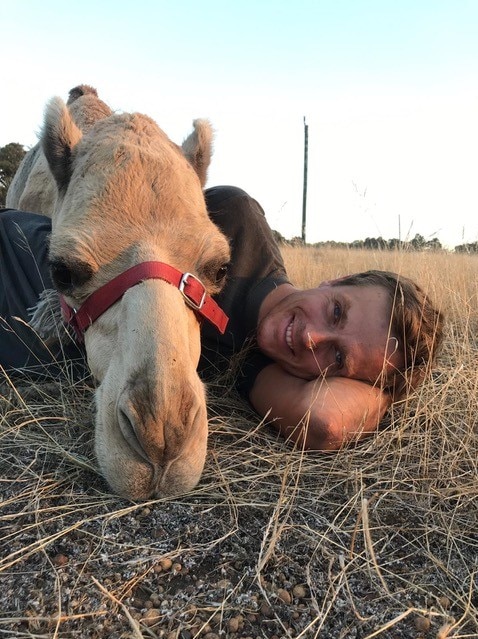 A man and camel lie on the ground looking at the camera.