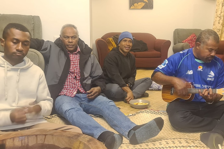 Four Fijian workers sit on the floor in a lounge-room, with one playing a ukele. 