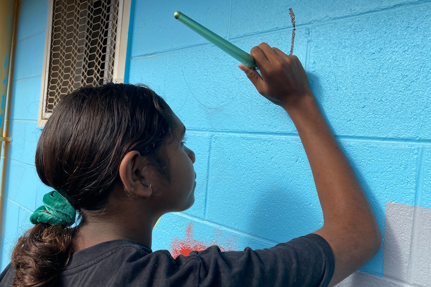 A profile view of an Aboriginal girl holding a paintbrush against a blue brick wall.
