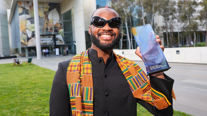 Genesis Owusu in shades and traditional dress holding up his ACT Young Australian of the Year trophy out on a lawn