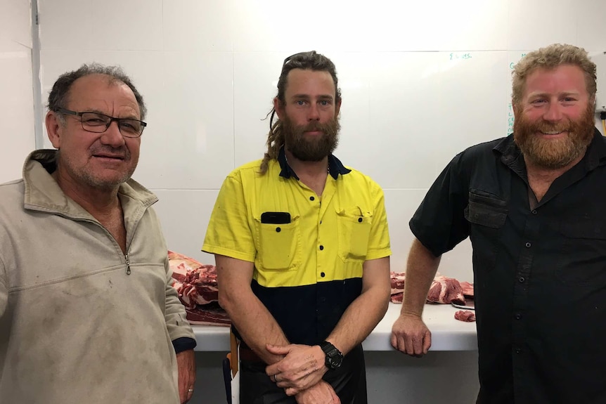Two farmers in work shirts flank younger man in high visibility clothing inside new, white butchers shop with meat in background