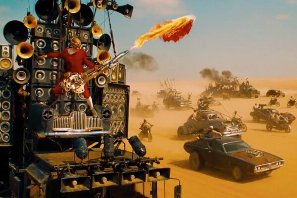 A man holds a guitar spouting flames while strapped to a vehicle in a desert filled with cars