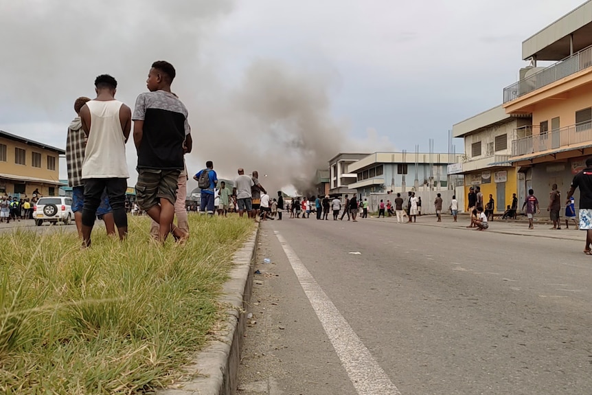 Crowds are seen on the streets of Honiara as smoke emerges from the distance