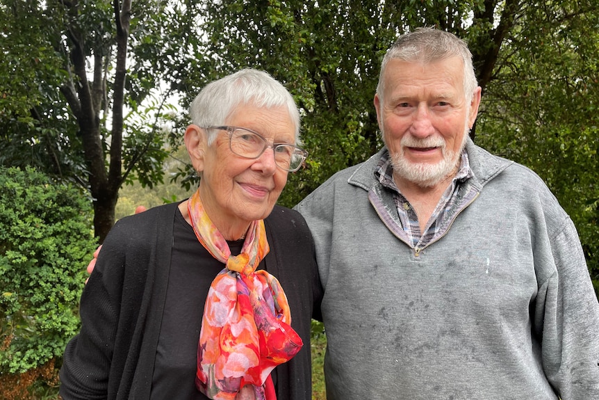 An older man and woman stand together with green trees behind them.