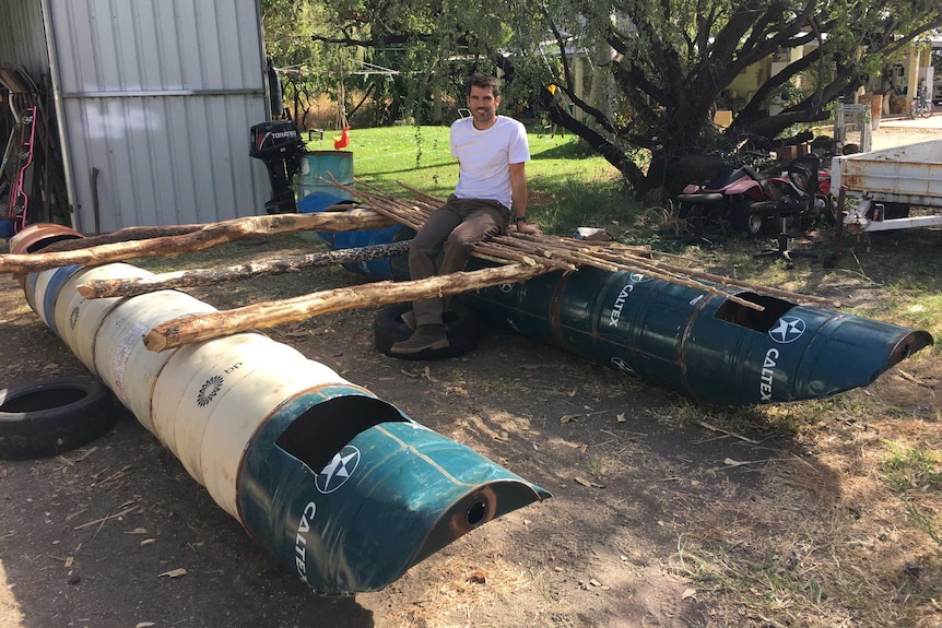 A man sits on a hand-made raft made from drums and timber in a yard.