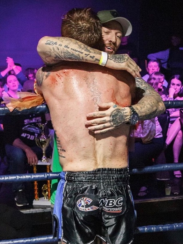 Two men, one shirtless, embrace in a kickboxing ring