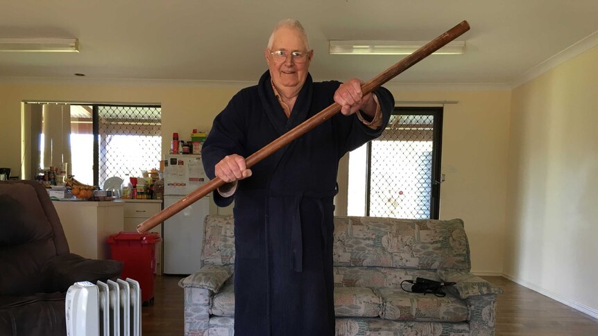 Elderly man in blue dressing gown stands in loungeroom holding large, wooden curtain rod.
