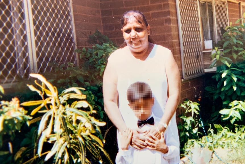 A woman photographed in front of a house with her arms around a small boy wearing a suit.