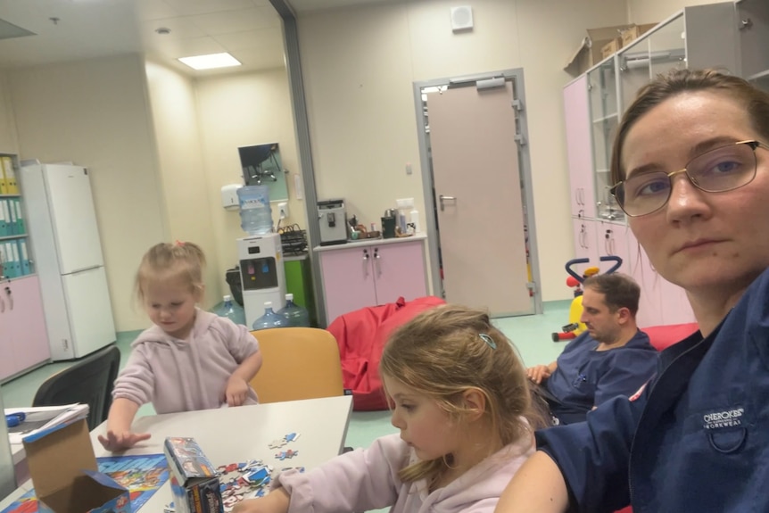 A woman in glasses and scrubs in the foreground as two young girls sit at a table drawing and a man in scrubs sits on the floor