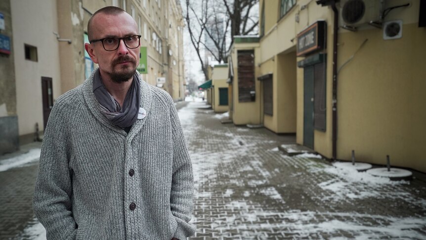  Konrad Korzeniowski stands in a snowy city street in a grey woolly jumper and glasses, with a neutral expression
