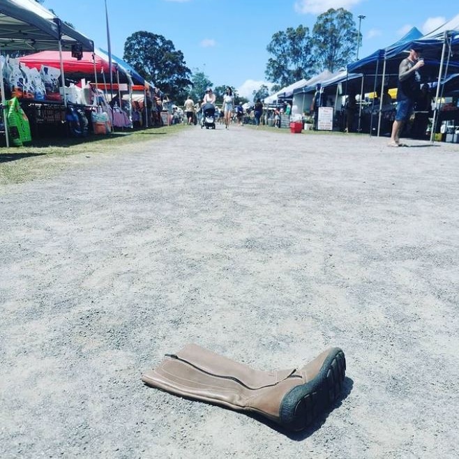 Knee-high brown boot lays on gravel path at an outdoor market, stalls and people walking in the background.