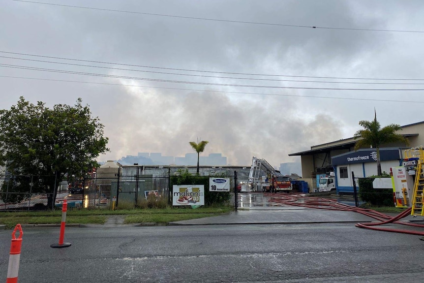 Smoke billows from a warehouse as two fire engines are parked at the scene. Road is wet from overnight rain.