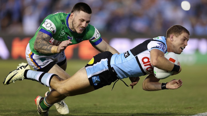 A Cronulla Sharks NRL player dives over the tryline with the ball as a Canberra defender tries to stop him.