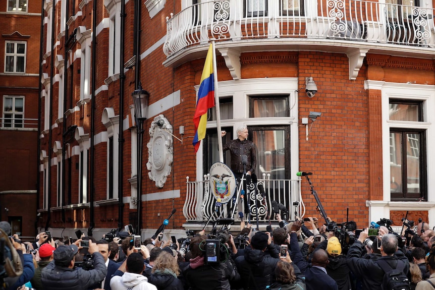 Assange raises his fist on the embassy's balcony in front of a large crowd of supporters and reporters with recording equipment.