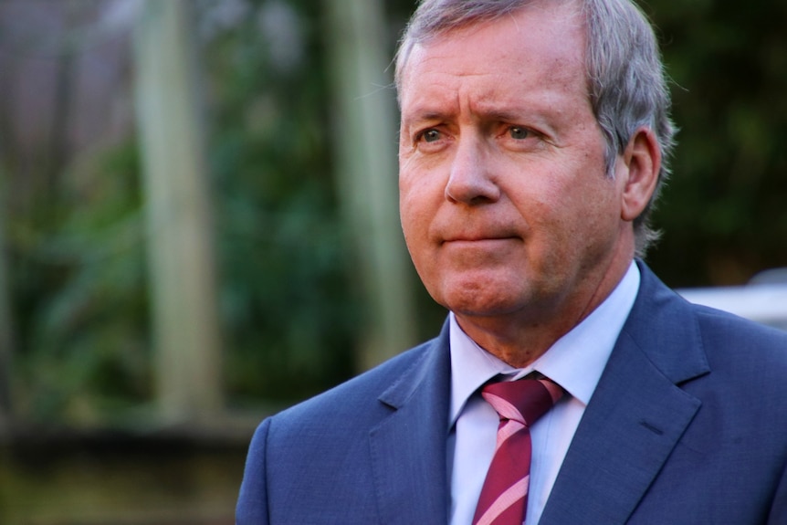 Mr Whitby stares into the middle distance. Wearing a blue suit and red tie.