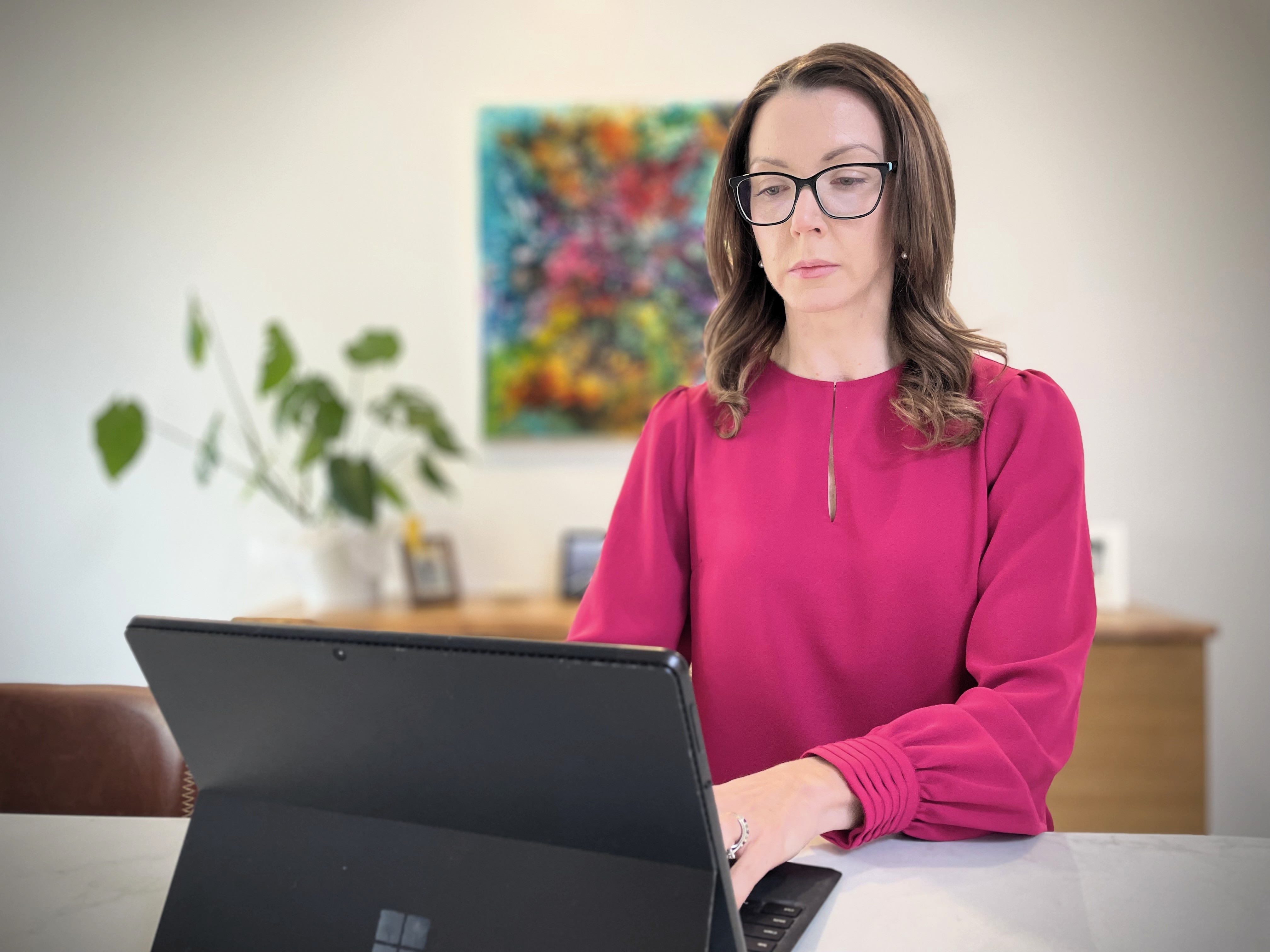 A white woman with long brown hair and glasses working on a laptop