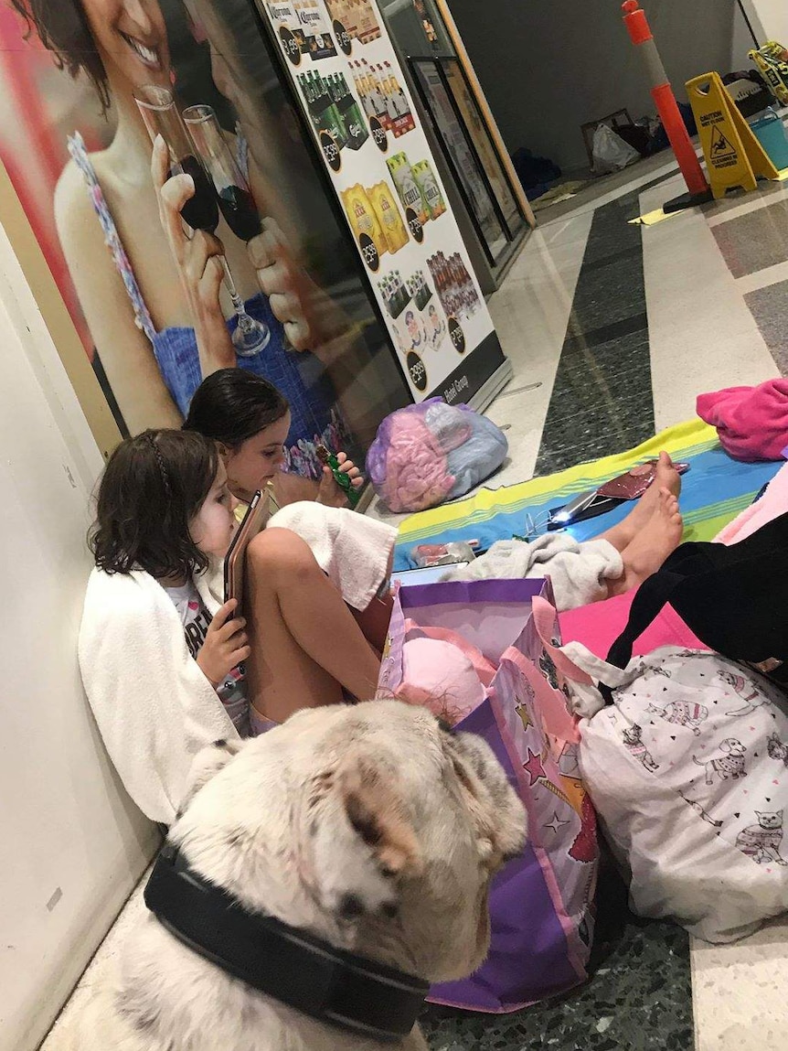 Two young girls sit on towels on a shopping centre floor with a white dog.
