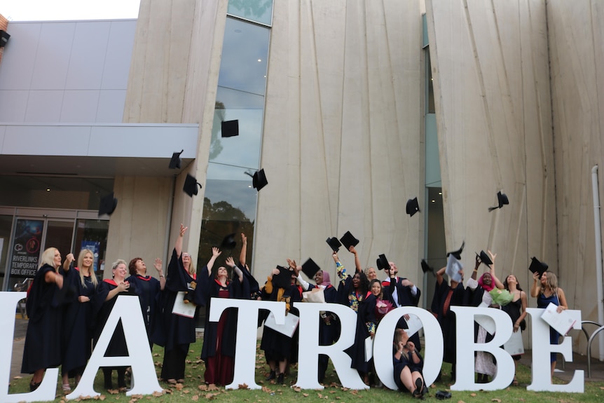 University students in graduation gowns throwing their hats in the hair in front of a sign that says La Trobe